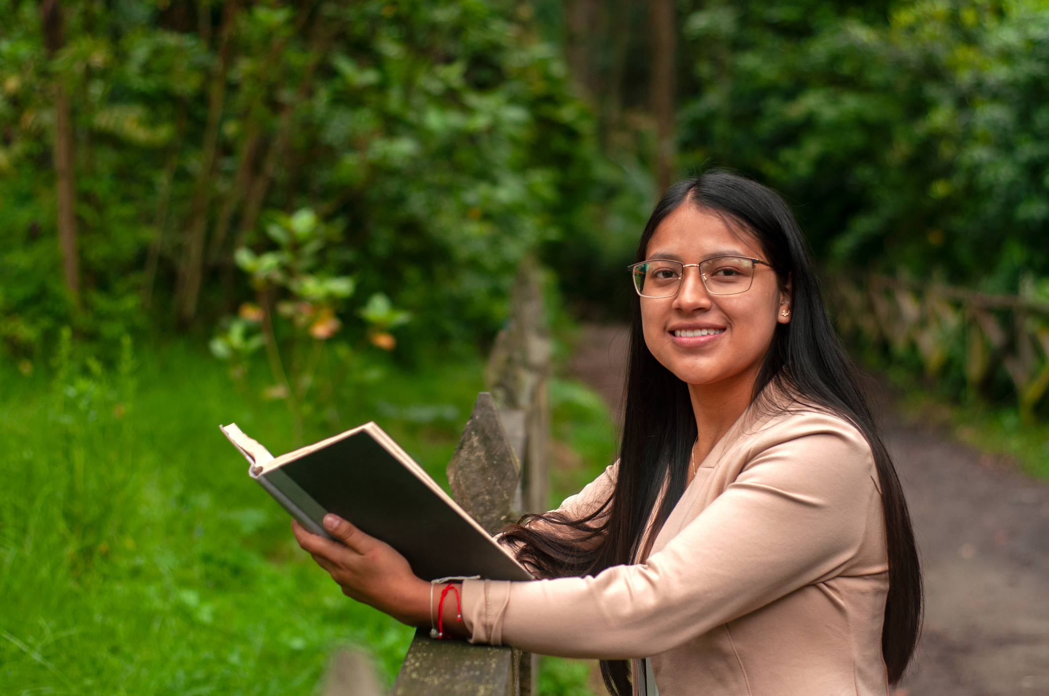 latina girl with glasses and long hair looking at the camera with a smile and an open book in her hands surrounded by greenery. book day